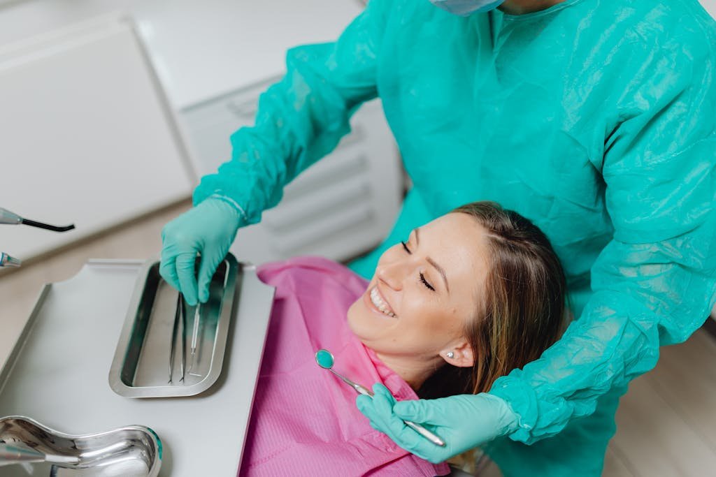 A woman sits smiling in a dental chair while a dentist prepares instruments.